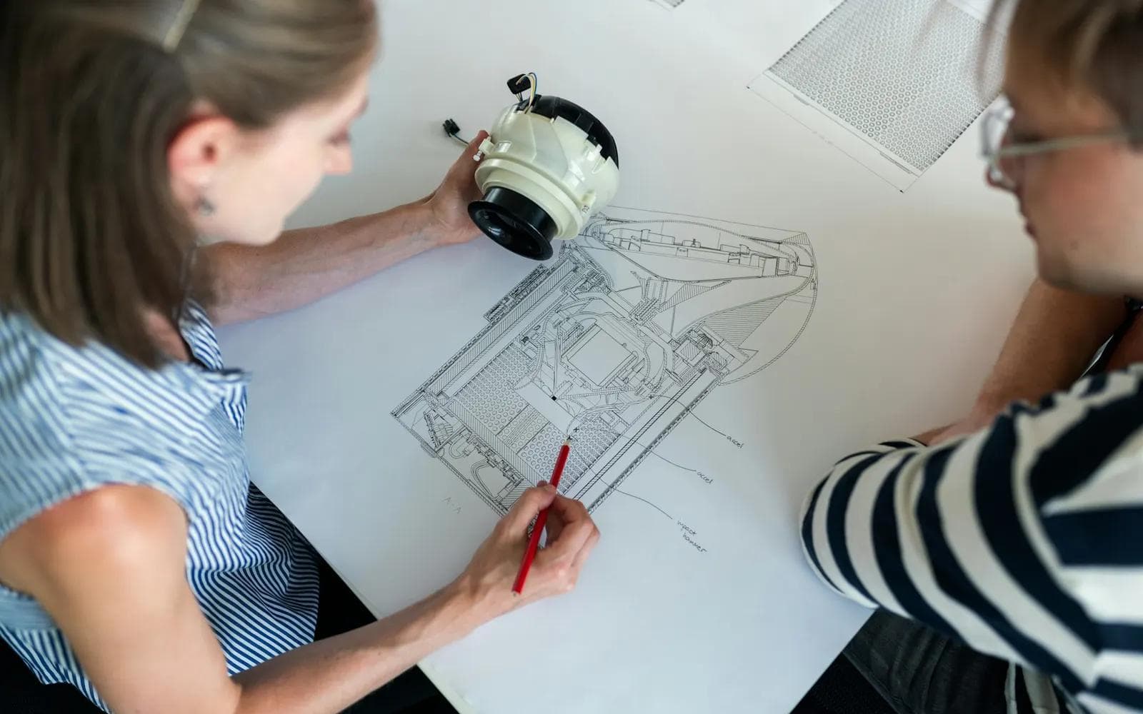 Pessoas analisando um desenho técnico e um componente, imagem ilustrativa